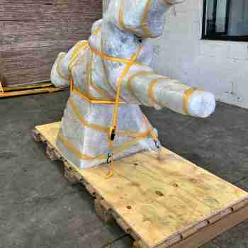 Reinforced honeycomb pallet securing a wrapped sculpture with straps for shipping.